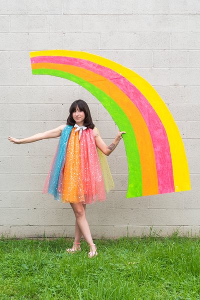 Marisol Muro photographed in her garden, wearing a rainbow-colored tulle mini dress. She is barefoot and holding a huge rainbow prop in her other hand.