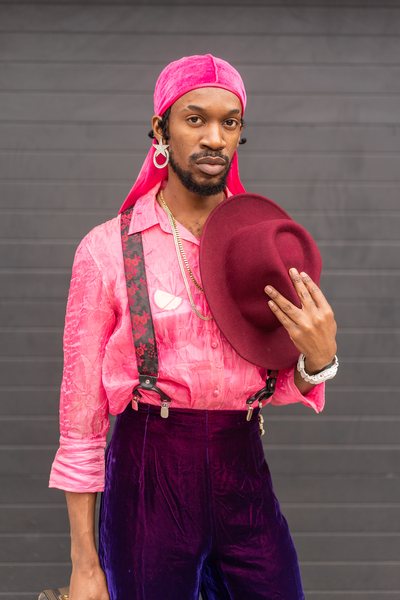 Kechi Onimo wearing a pink velvet durag, a pink see-through thrifted shirt, suspennders, and velvet thrifted pants. In front of his chest he is holding a red hat.
