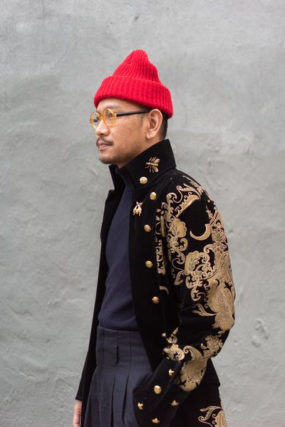 Illustration artist Prates Songtieng, 35, also known as Monsieur Poppi, standing in front of a grey wall and wearing a red beanie, orange frames, dark blue turtleneck and richly embroidered high collared navy and gold vintage jacket