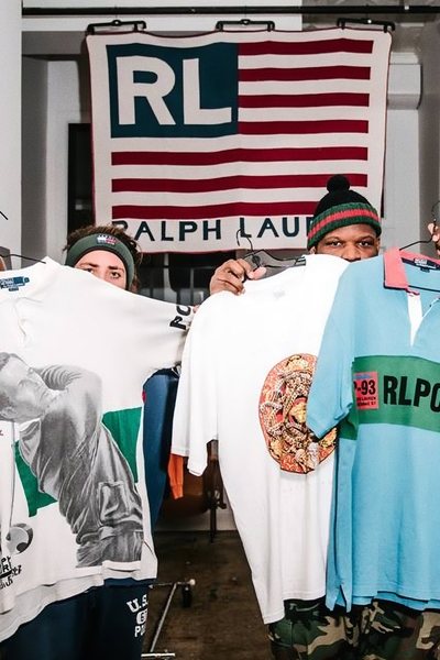 Vintage seller Mark Rosado from Top Shelf Premium and another person holding three different Polo Ralph Lauren t-shirts in front of a big Ralph Lauren canvas