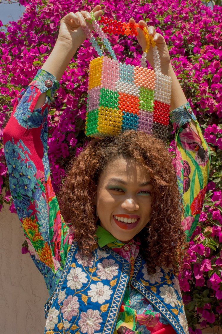 A smiling girl holding a beaded Susan Alexandra style handbag on top of head, wearing a colorful floral button up and blue embroidered floral vest 