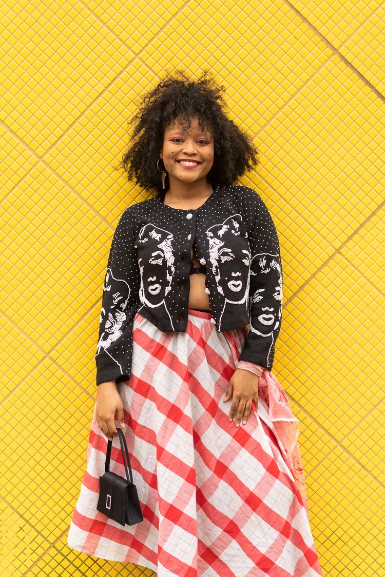 NYC-based queer artist Grey... smiling in front of a yellow tile wall and wearing a 50s black cardigan with embroidered Marilyn Monroe figures, an a-line red-and-white cotton skirt, and a black box shaped mini purse