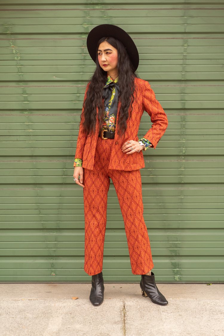 a woman wearing a black fedora hat, orange 60s 70s vintage suit, and a colorful vintage print blouse.