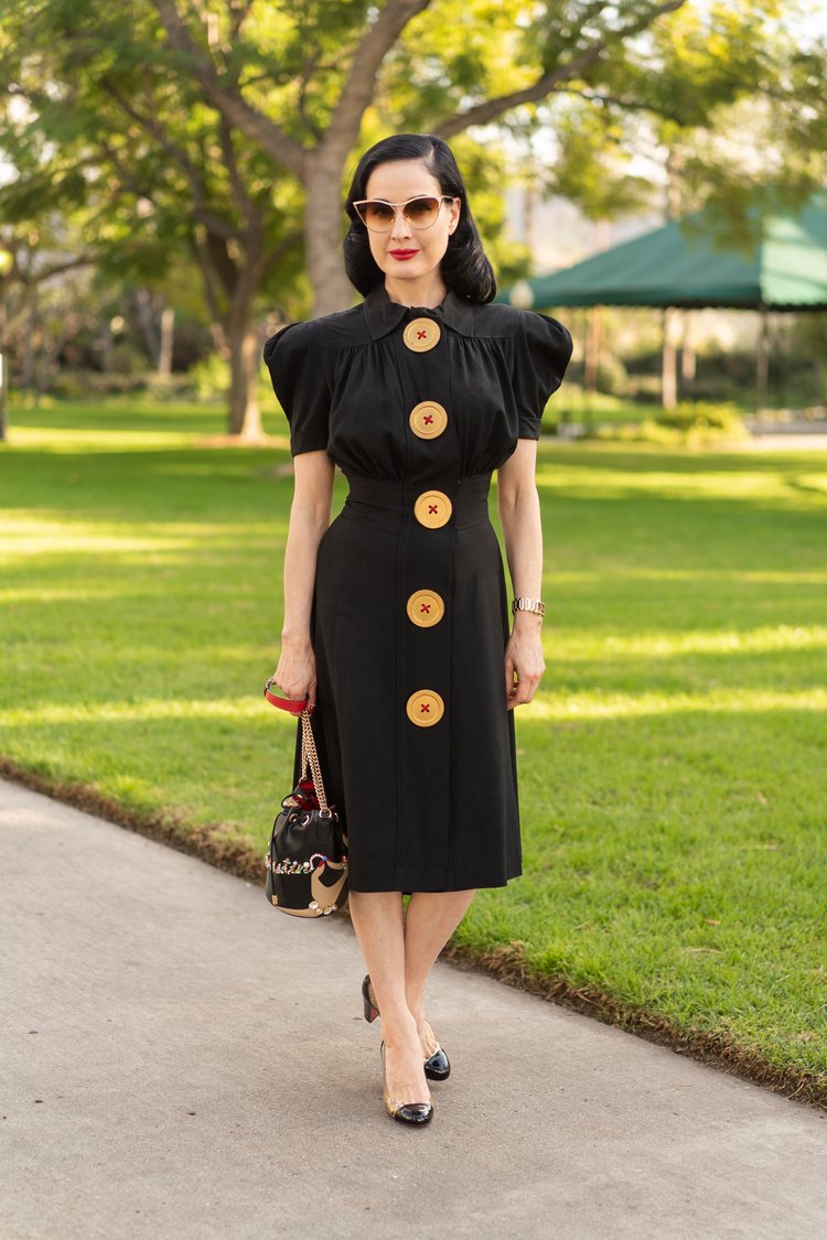 Dita von Teese in a 40s style black dress that has huge buttons and shoulder pads, cat eye sunglasses, and Chanel shoes. 