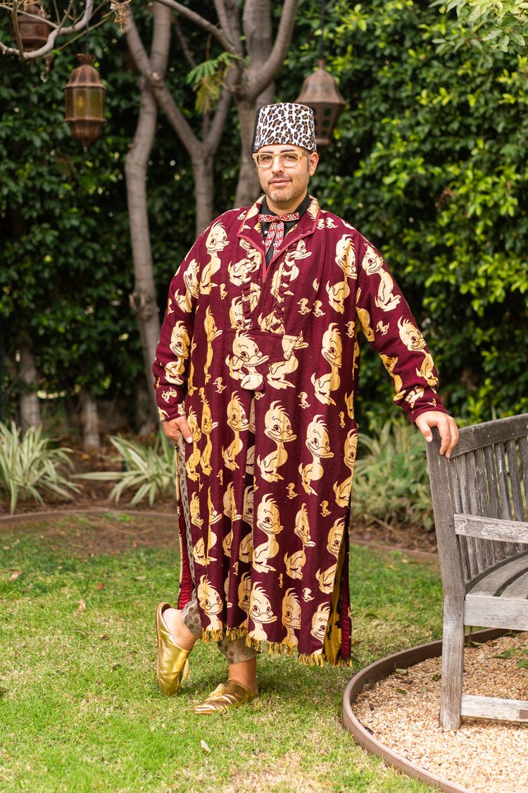 Ari Seth Cohen standing in his garden and wearing a golden and red velvet-t caftan with a leopard printed hat and golden slippers