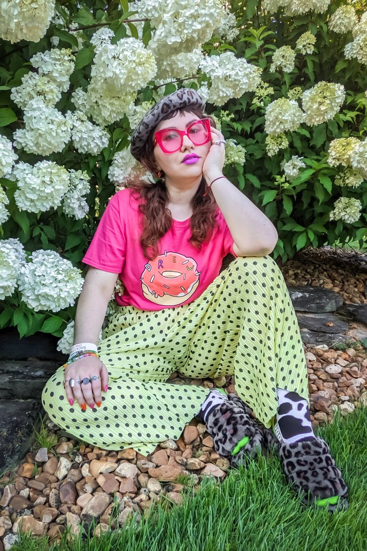 Sarah Stepanek sits on the ground in front of a blooming hydrangea bush. She is wearing a thrifted black hat from ThredUp, thrifted pink print Drew T-shirt from ThredUp, pleated pale yellow pants with black dots, UGGs shoes, socks from hop Mad Cool and a flower bead ring from Planet Solana