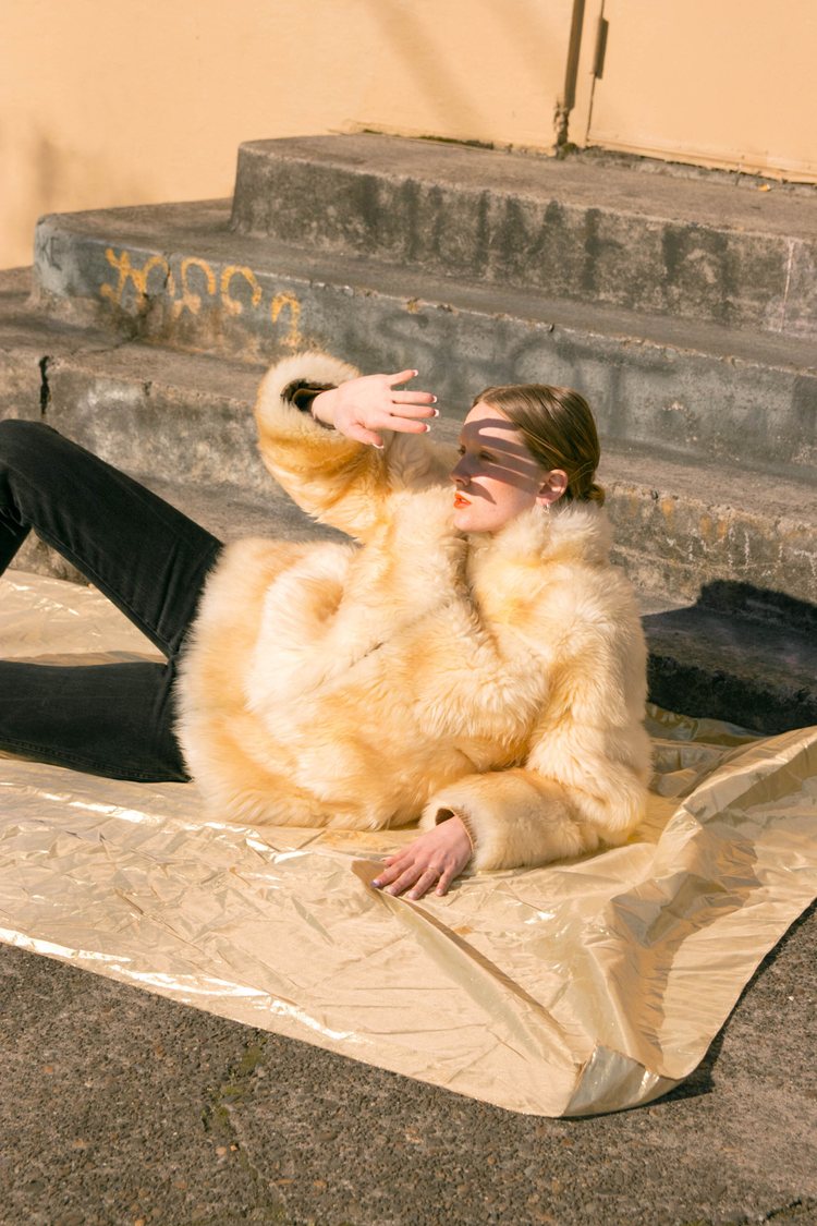 A model laying on the ground and looking towards the sun, wearing a vintage fur coat and black pants.