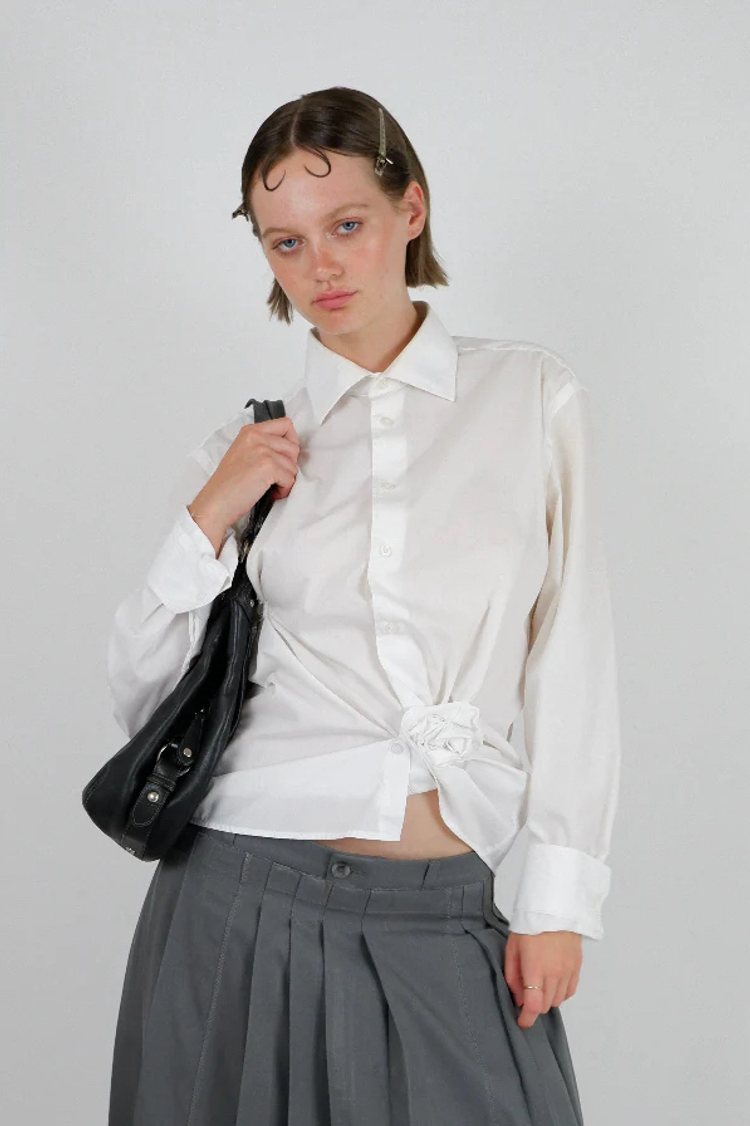 a young woman wearing a white button up vintage shirt with a rosette decoration, a grey pleated skirt, and a black shoulder bag