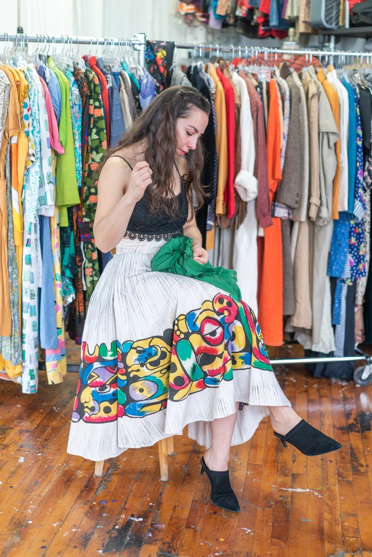 vintage seller Naomi Bergknoff sitting on a stool in her studio, mending a green vintage garment