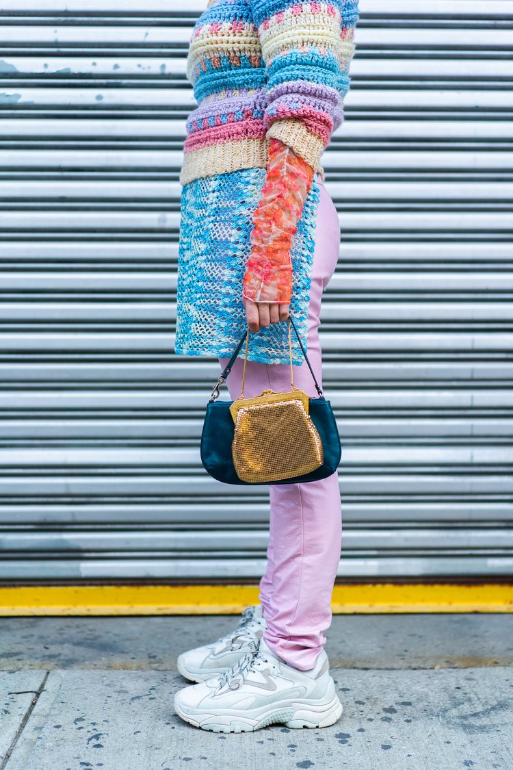 a layered pastel colored outfit consisting of a striped long sleeve crochet sweater, pink leather trousers and a blue crochet apron. The model has two mini bags in her hand, one is a golden mesh bag and the other one is navy blue leather bag.