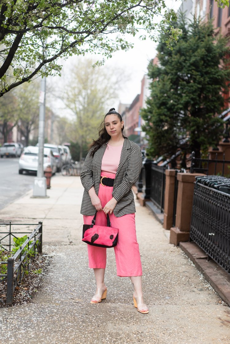 founder of Shop Berriez, Emma Zack, standing on the sidewalk in a black-and-white houndstooth vintage blazer, pink shirt, pink cropped linen pants, pink handbag, black wide belt and see-through slingback mule sandals