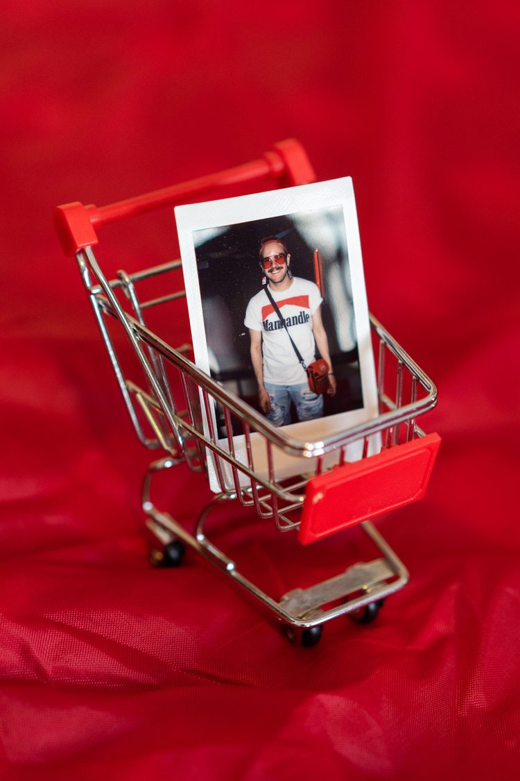 tiny toy supermarket shopping cart on a red fabric. Inside the cart, there is a Polaroid photo of a man wearing a white vintage Marlboro t-shirt