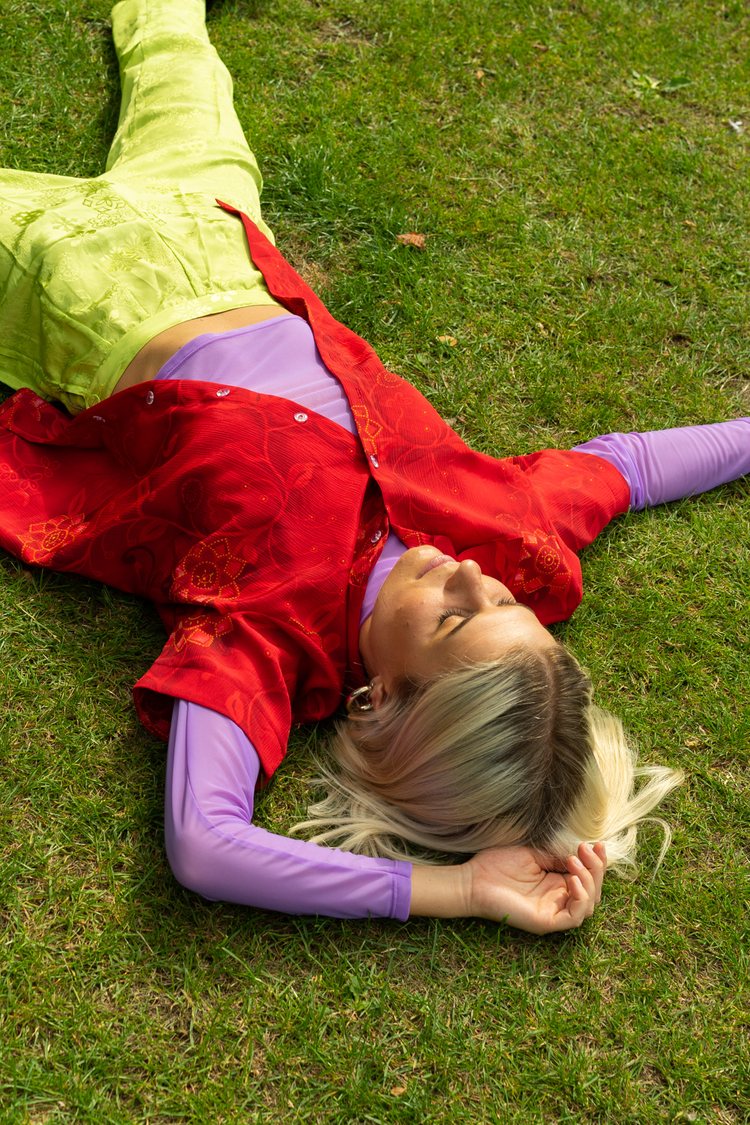 a young woman laying on the grass and wearing a colorful secondhand outfit: a purple mesh 90s long sleeve top, bright red button down and green-yellow shell suit pants